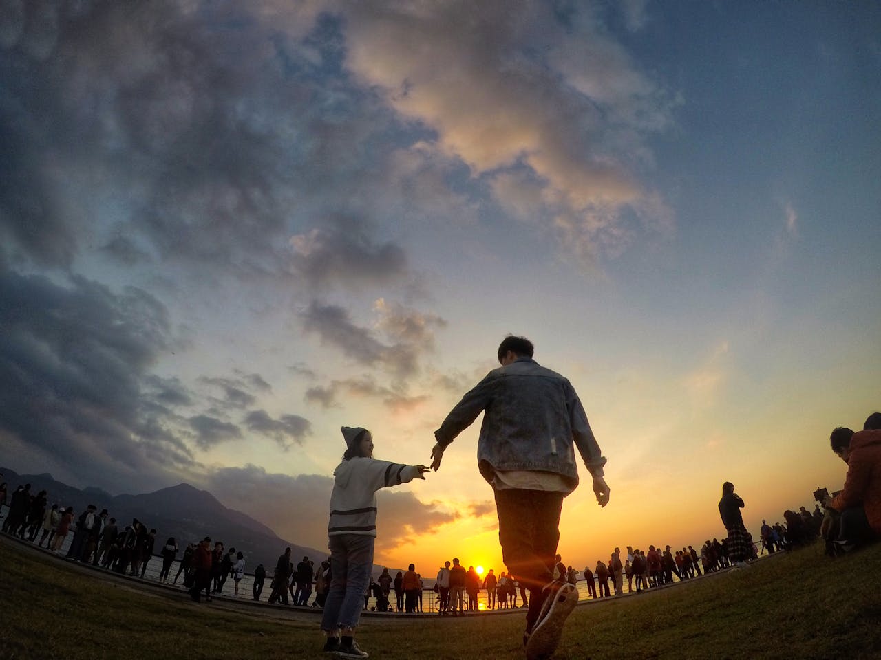 A couple holding hands silhouetted against a vibrant sunset sky with a crowd in the background.